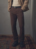 Person wearing a brown jacket, beige sweater, and brown pants standing on a patterned rug.