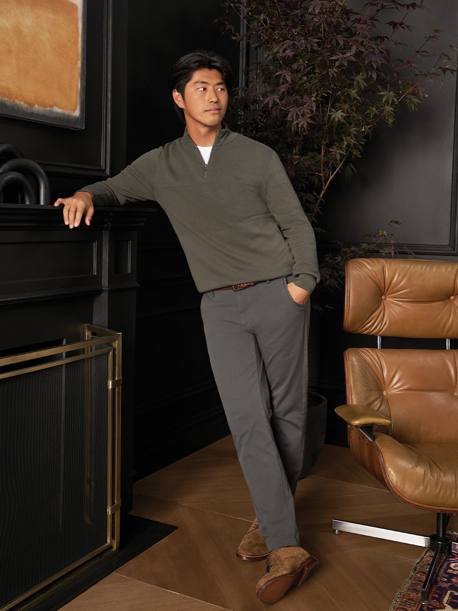 Man in a green sweater and gray pants standing in a room with a brown leather chair and black fireplace.