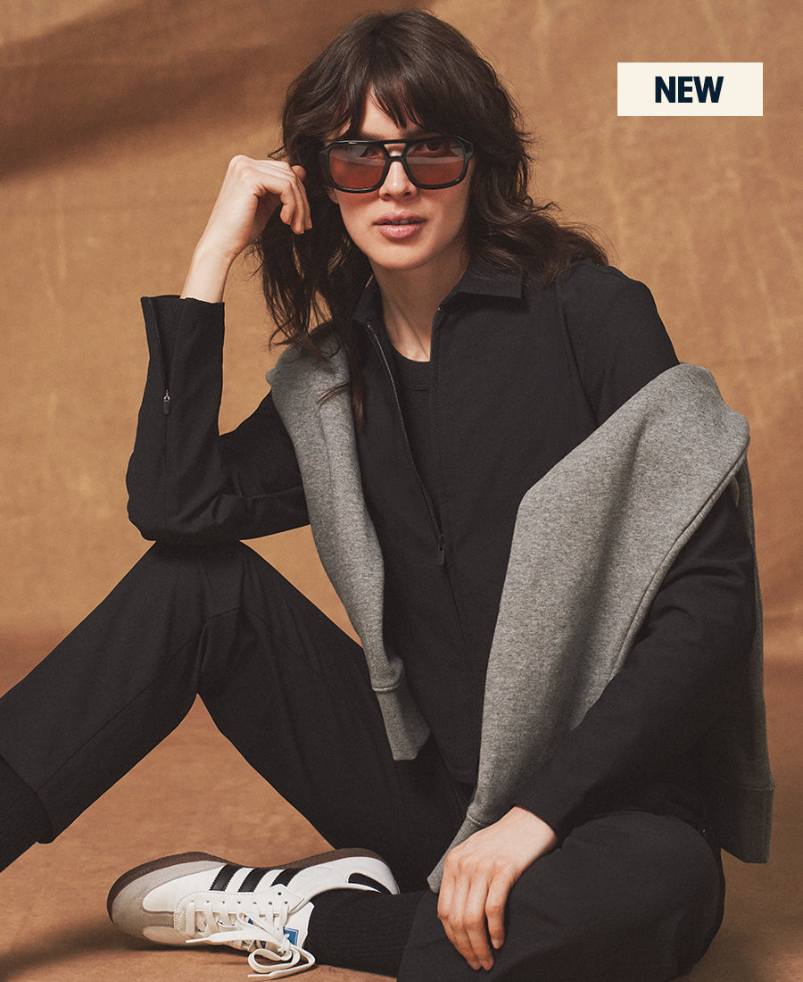 Person wearing sunglasses and a black outfit with a gray sweater draped over their shoulders on a brown background.