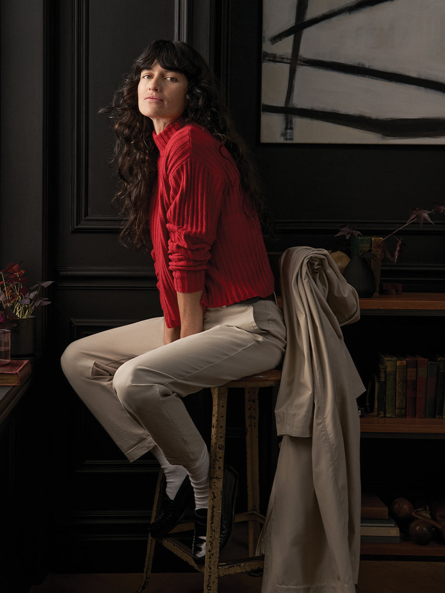 Woman in a red sweater sitting on a stool in a room with books and a painting.